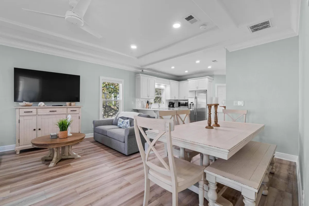 This image showcases a spacious, sunlit living room with a large, comfortable sofa, a dining table perfect for group meals or games, a prominently placed smart TV for movie nights, and an adjacent kitchen area. The open-concept design allows for seamless movement and interaction between spaces, ideal for gatherings and relaxation after a day at the beach.