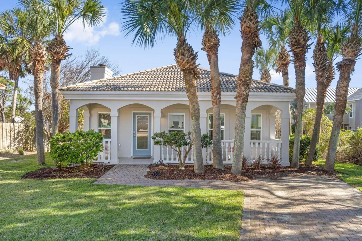 The image showcases the charming exterior of a Destin beach cottage, featuring a welcoming front porch with white railings and graceful archways, a striking blue front door, and tall palm trees lining a brick-paved walkway leading to the entrance. The atmosphere is peaceful and inviting, hinting at the coastal lifestyle and comfort awaiting inside.