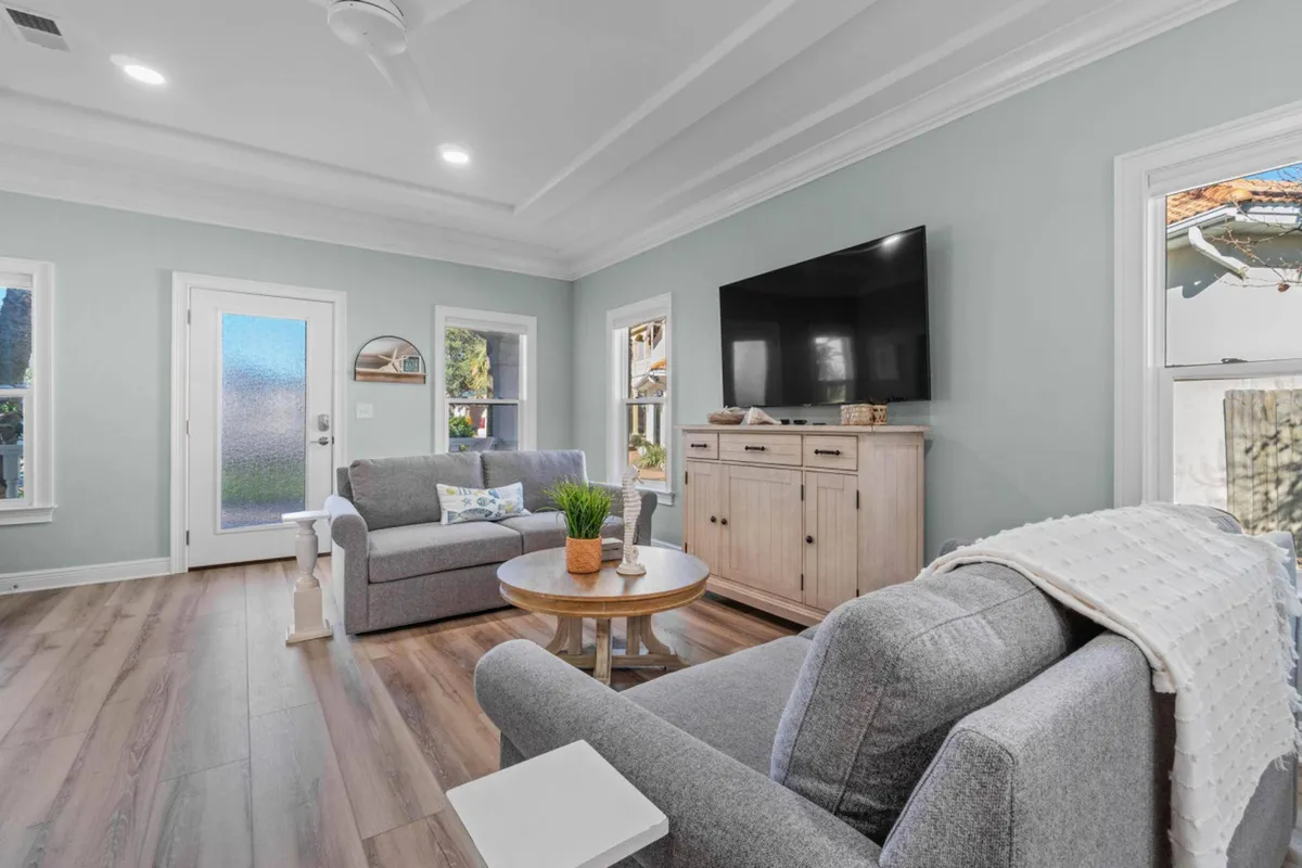 A bright, welcoming living room with two gray sofas facing each other, a round wooden coffee table with decorative items, a light wood TV cabinet with a mounted flat-screen TV, and large windows that let in abundant natural light. The space radiates comfort and encourages relaxation or group conversation.