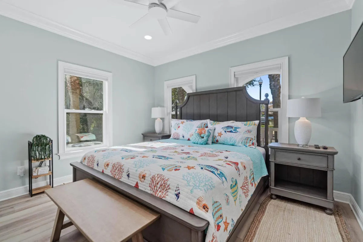 A bright, airy master bedroom featuring a king-sized bed adorned with an ocean-themed quilt. Three windows fill the space with soft natural light, while matching wooden nightstands with lamps and a wall-mounted TV create a cozy, restful environment ideal for relaxation after a day in the sun.