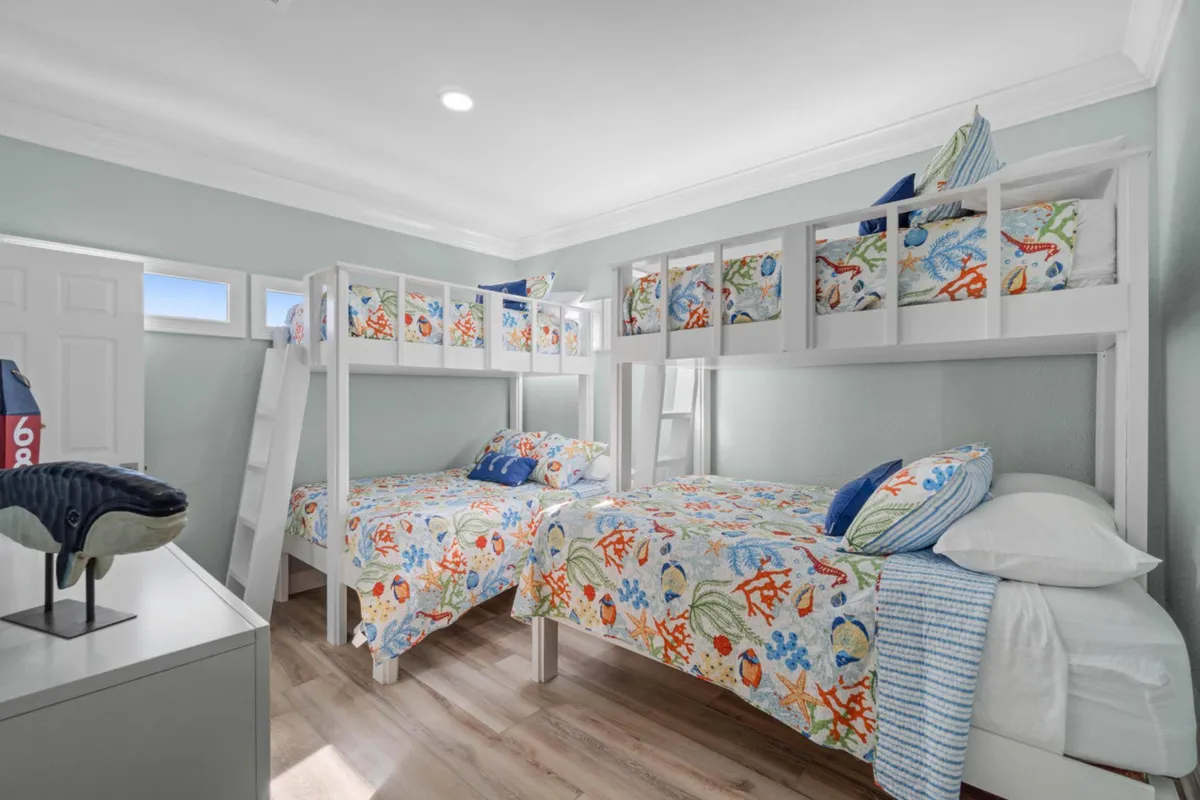A cheerful second bedroom with two white wooden bunk beds featuring colorful sea-themed bedding. The space is bathed in bright natural light from windows, includes a whale figurine on the dresser, and has light wood-look flooring—making it playful and inviting for children or groups.