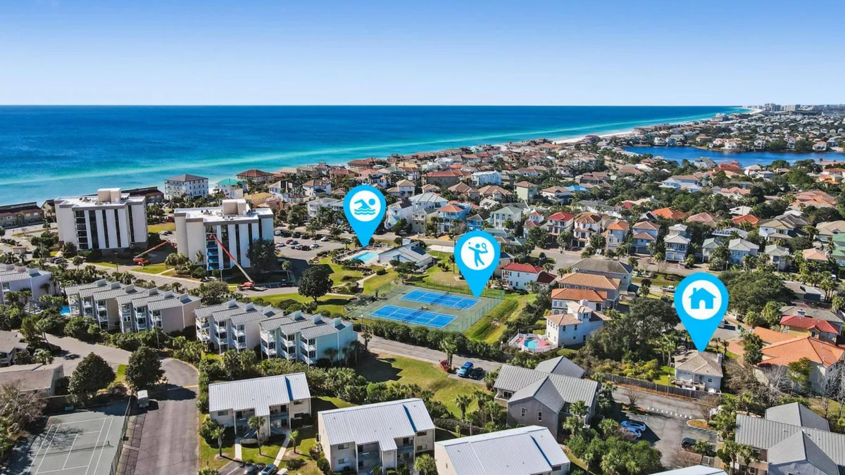 An aerial view captures both the private beach access near the coastline and the sparkling community pool, along with tennis courts and a cluster of cozy residential homes. The scene exudes a sense of resort convenience, beach proximity, and recreational amenities.