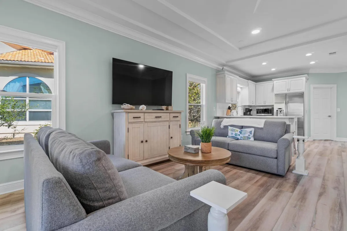 A comfortable living space with gray sofas arranged for conversation, a large wall-mounted flat-screen TV, a wooden coffee table with decorative plant and books, and an open kitchen with white cabinetry and stainless steel appliances in the background. Natural light enhances the relaxed, inviting mood.