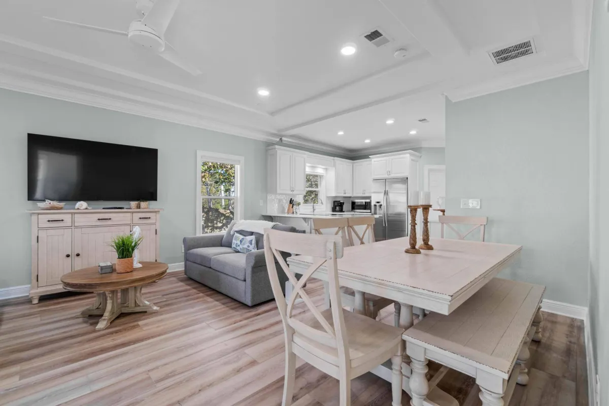 A comfortable, stylish interior space featuring a dining table with bench seating, a plush grey sofa adorned with decorative pillows, a wooden coffee table with a plant centerpiece, and a modern kitchen with stainless steel appliances. The layout encourages togetherness, relaxation, and easy living after a day at the beach or pool.
