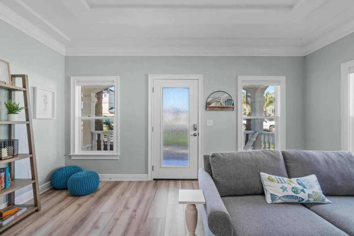 A cozy living space featuring a gray upholstered sofa with a coastal-themed pillow, textured blue poufs positioned by a sunlit window, a decorative ladder shelf with books and plants, and a door with a frosted glass panel leading outside. The decor and layout suggest easy access to outdoor adventures and a relaxing place to return after a day at the beach or pool.