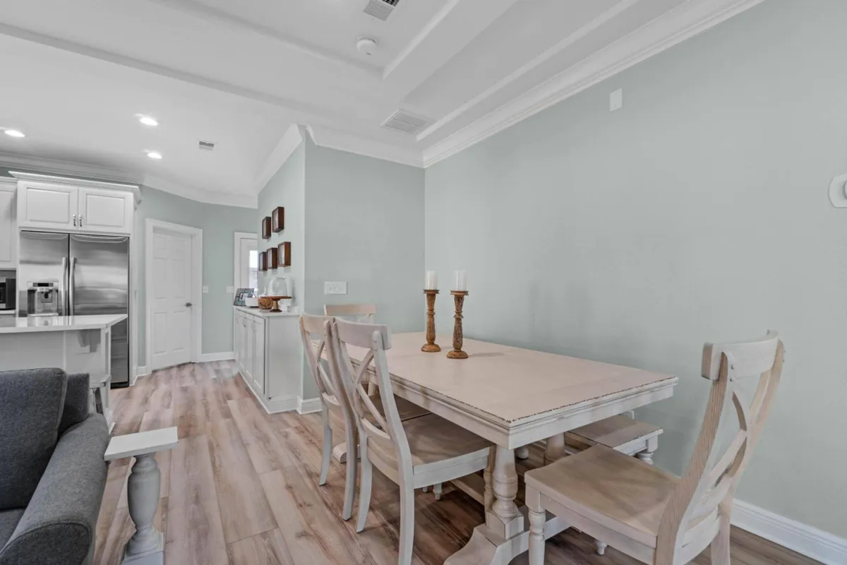 A bright and welcoming dining area featuring a wooden dining table with chairs, set for up to eight guests. The table is adorned with candlesticks, and in the background, the kitchen area with stainless steel appliances is visible. Light wood flooring enhances the coastal, airy atmosphere, and the adjacent living area is partially visible, creating a sense of open-plan living.