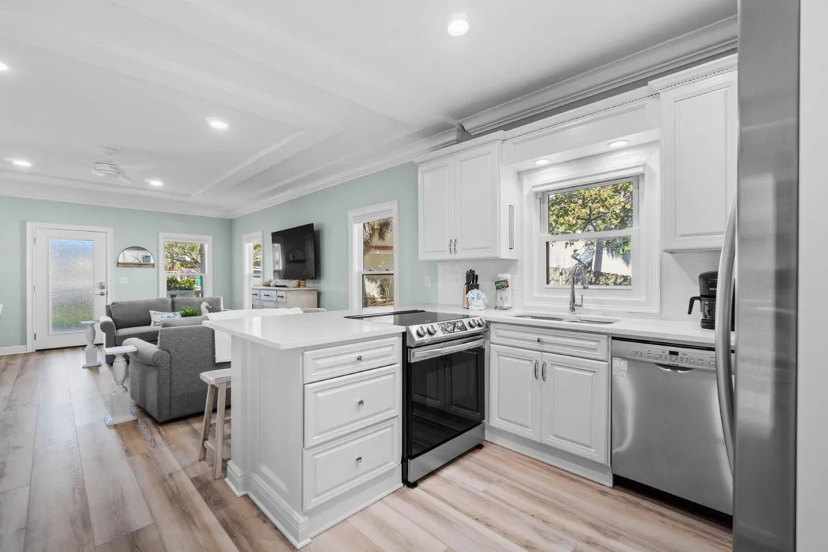 A modern kitchen with white cabinetry, quartz countertops, and stainless steel appliances such as a stove and dishwasher. The kitchen opens into a living area with a mounted flat-screen TV and comfortable seating, with light wood flooring tying the spaces together. The overall setting is bright, fresh, and conducive to cooking and socializing.