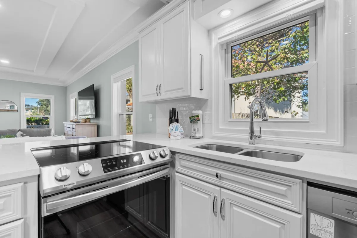 A fresh, welcoming kitchen scene with a stainless steel electric stove and oven, a double kitchen sink under a window, white cabinetry, and an open view into the living room with a TV and sofa. The setup suggests easy movement between cooking, relaxing, and socializing.