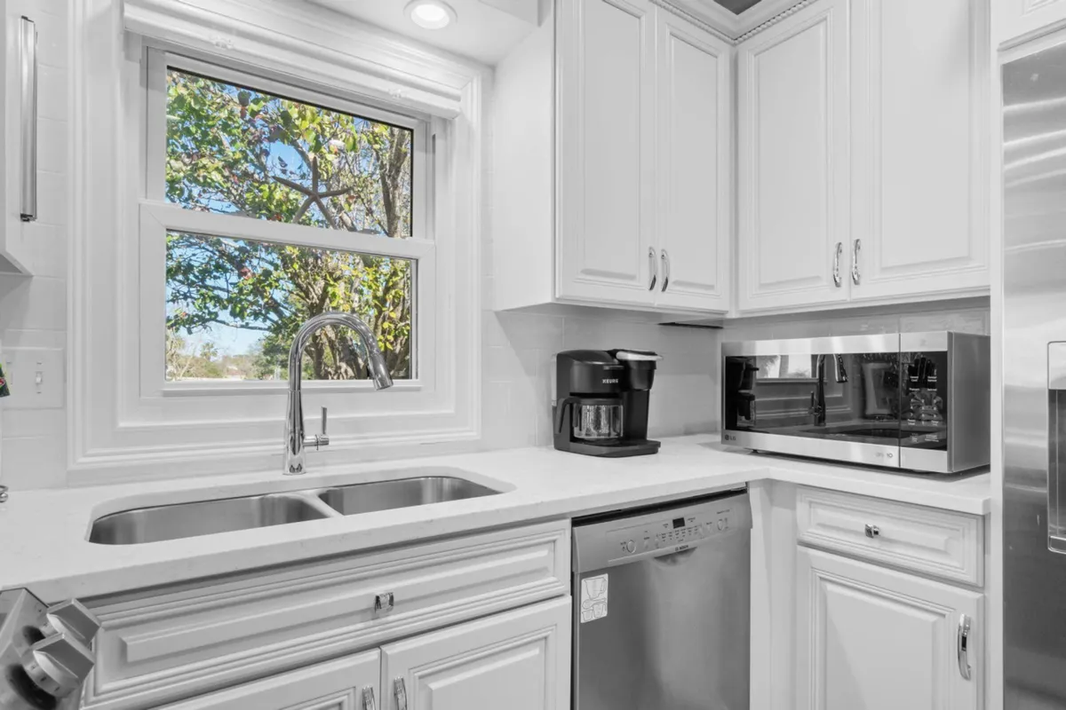 A bright, modern kitchen with a double sink under a window, stainless steel dishwasher, black coffee maker, and microwave oven. The scene is filled with natural light, and the window provides a glimpse of greenery outside. The kitchen feels welcoming and is set up for both meal preparation and casual coffee breaks, with modern amenities in a cozy cottage setting.