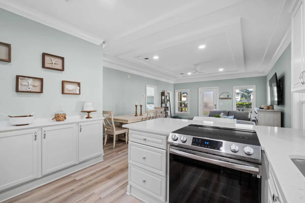 A bright, open-plan kitchen and living area featuring a kitchen island with a stainless steel stove, a dining table with chairs set for a group meal, and a welcoming seating area with a TV. Coastal wall art and large windows create a breezy, inviting atmosphere ideal for socializing and group gatherings.