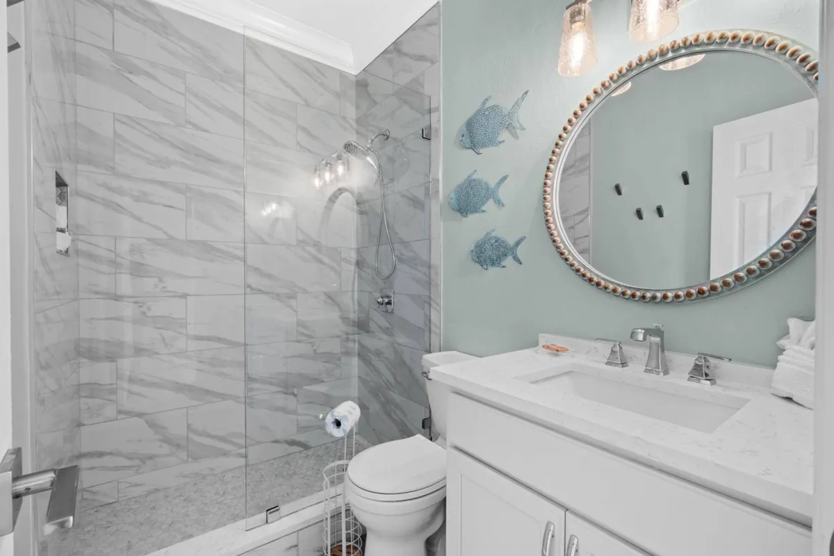 A modern bathroom with a marble-tiled walk-in shower enclosed by a glass door, a round decorative mirror above a white vanity, and subtle coastal decor like metal fish wall art. The space feels refreshing and calming—ideal for spa-like winddowns.