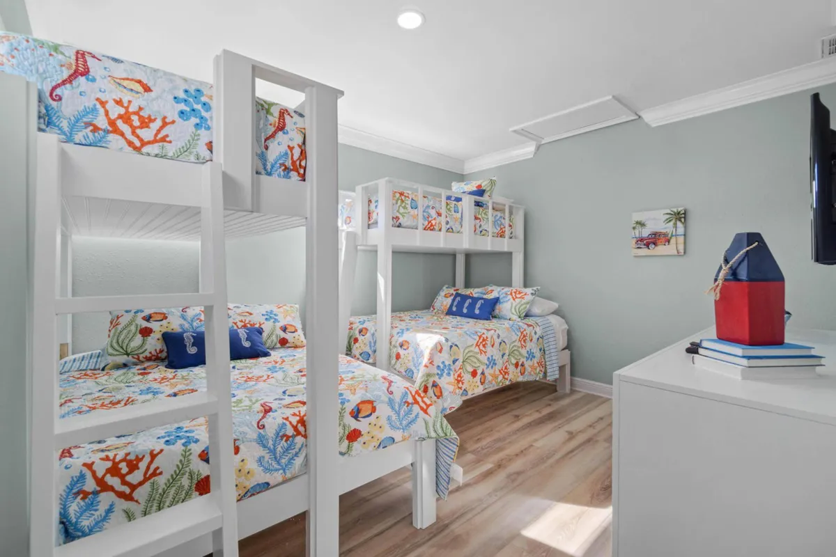A cheerful children's room with two white bunk beds dressed in colorful sea-themed bedding, light wood flooring, a wall-mounted TV, and nautical-themed decor atop a dresser. The room is bright, playful, and tailor-made for family adventures.