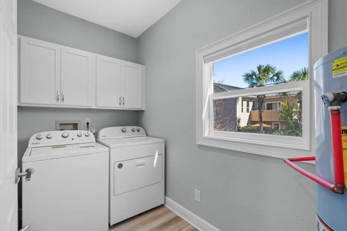 A bright laundry room featuring a white top-loading washing machine, matching dryer, and a window looking out onto palm trees and a neighboring house. Wall cabinets above the appliances keep the space organized, and the window bathes the room in natural light, creating a sense of openness and connection to the surroundings.