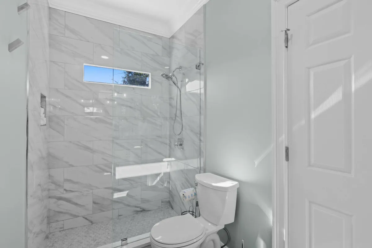 A clean, modern bathroom featuring a glass-enclosed shower with marble tile, a toilet, and a small horizontal window that brings in natural light, creating a bright and fresh atmosphere.
