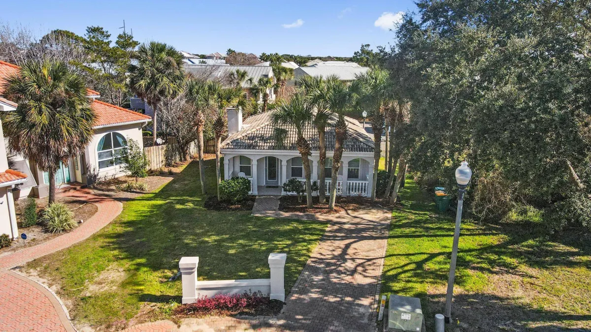 This exterior shot features a charming coastal cottage with a welcoming front porch, surrounded by lush palm trees and a brick walkway. Neighboring homes with similar architecture are visible, emphasizing a tranquil, community-oriented setting. The inviting porch and greenery underscore the pet-friendly, leisurely lifestyle promoted in the section.